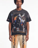 A man wearing a faded black graphic t-shirt featuring a muscular bulldog in a wrestling singlet, paired with beige Allstar pants with orange graphics, against a plain white background.