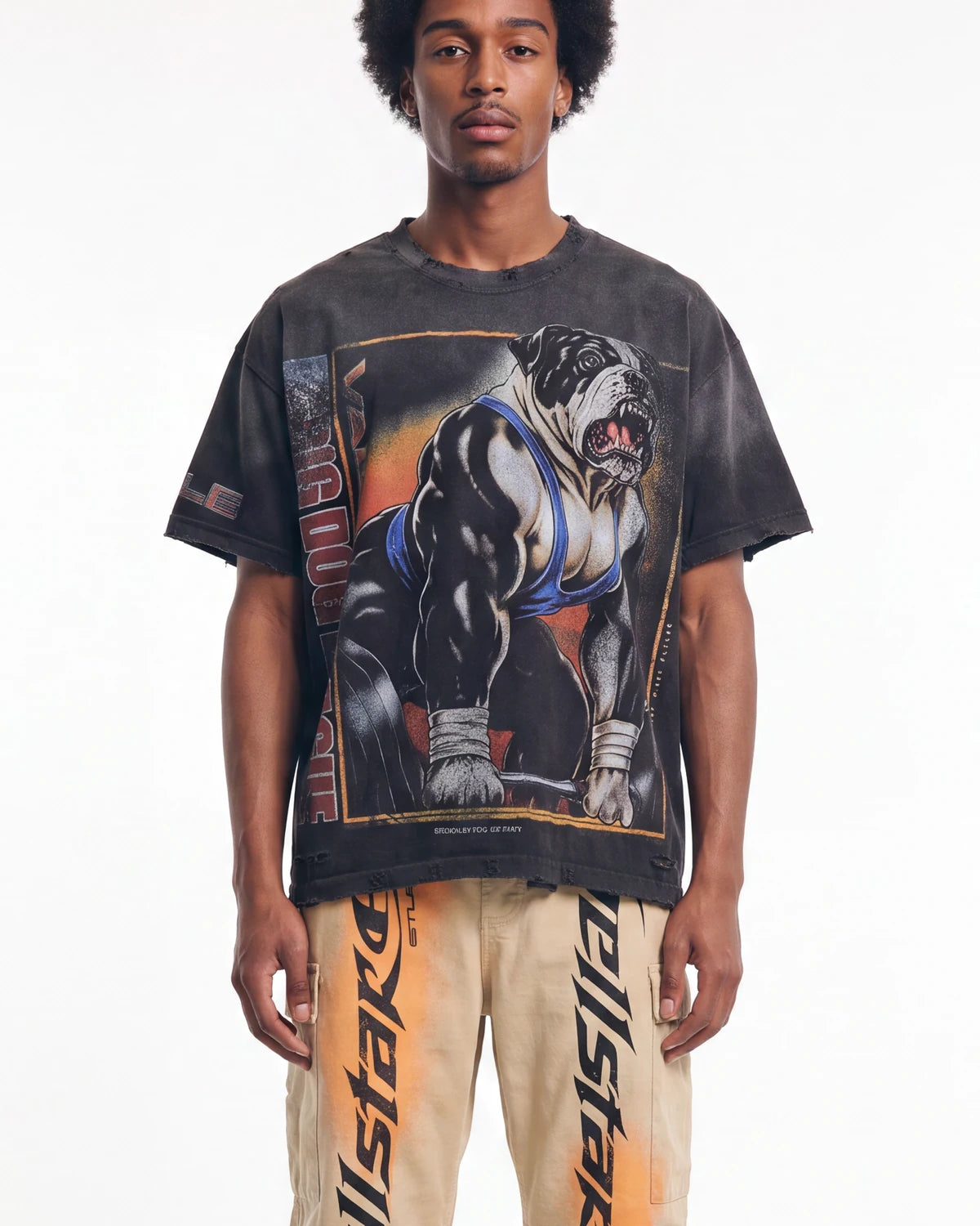 A man wearing a faded black graphic t-shirt featuring a muscular bulldog in a wrestling singlet, paired with beige Allstar pants with orange graphics, against a plain white background.