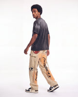 A man wearing beige cargo pants with orange and black graphics, a faded black t-shirt, and sneakers, shown from a side-back angle against a white studio background.
