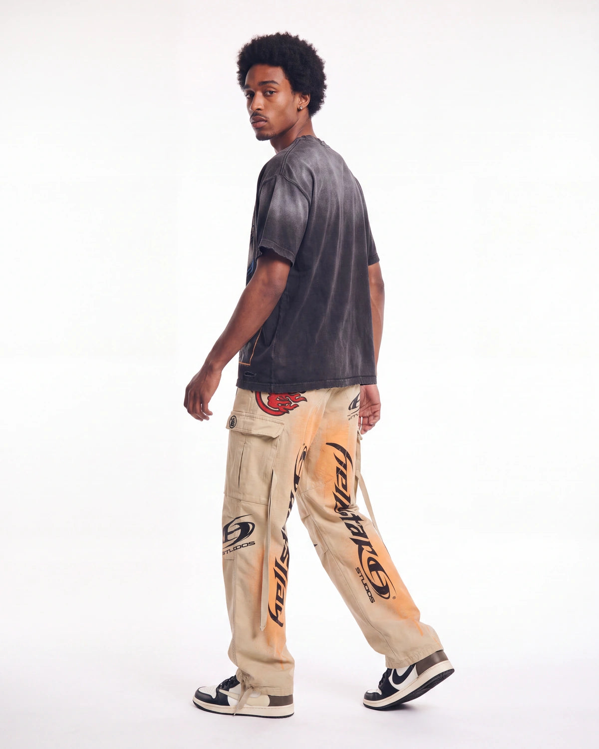 A man wearing beige cargo pants with orange and black graphics, a faded black t-shirt, and sneakers, shown from a side-back angle against a white studio background.