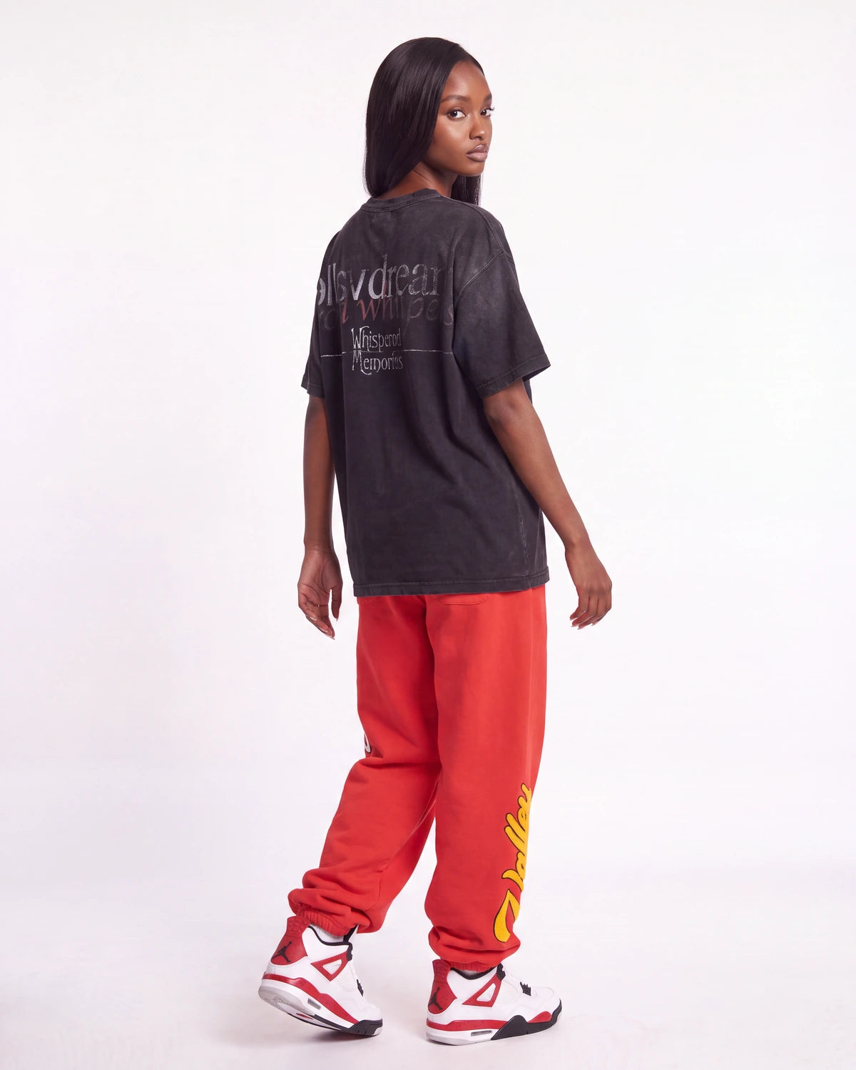 A model shown from the back wearing a faded black graphic t-shirt, bright red sweatpants with yellow graphics, and white sneakers with red accents, set against a plain white studio background.