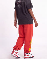 A person shown from the back wearing bright red sweatpants with a large yellow graphic print on the side, a faded black graphic T-shirt, and white sneakers with red and black accents, against a plain white studio background.