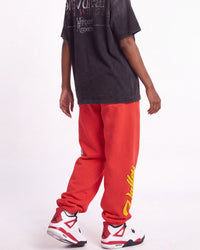A person shown from the back wearing bright red sweatpants with a large yellow graphic print on the side, a faded black graphic T-shirt, and white sneakers with red and black accents, against a plain white studio background.