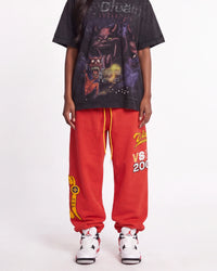 A person wearing an oversized black graphic T-shirt, bright red sweatpants with yellow and white graphics, and white sneakers, standing against a light gray studio background.