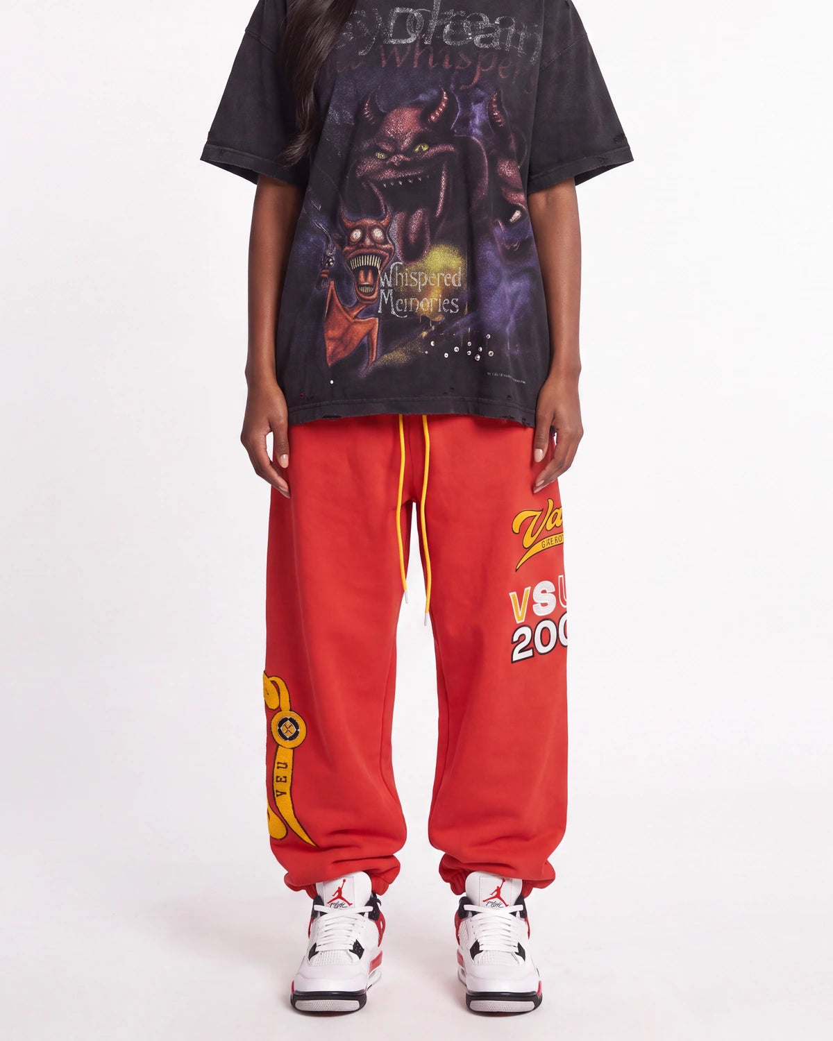 A person wearing an oversized black graphic T-shirt, bright red sweatpants with yellow and white graphics, and white sneakers, standing against a light gray studio background.
