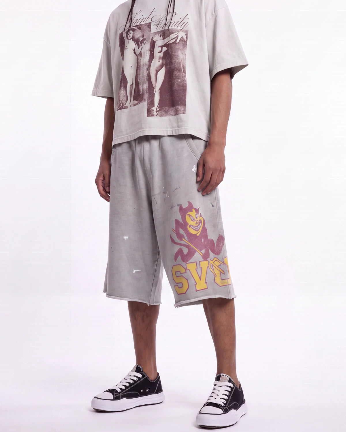 A person modeling a light gray graphic t-shirt and matching light gray distressed sweat shorts with a purple and yellow graphic on the left leg, paired with black low-top sneakers, against a plain white studio background.