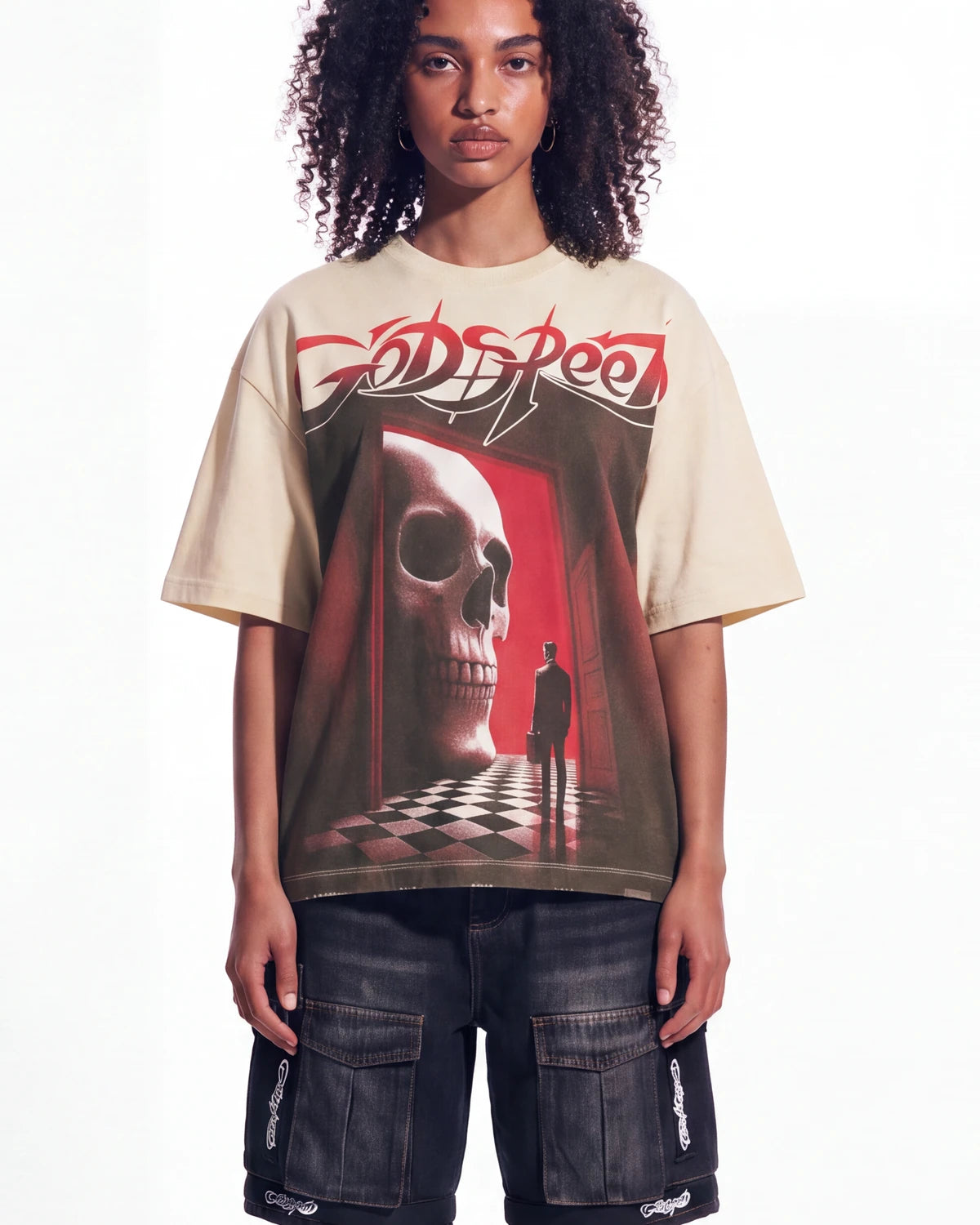 A model wearing a beige Godspeed t-shirt with a large red and black graphic print of a skull in a hallway, paired with dark cargo shorts, against a plain white background.