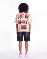 A child model shown from the back wearing a cream-colored t-shirt with bold red and black text reading 'OUT OF MANY, ONE' and dark denim shorts, against a plain white studio background.