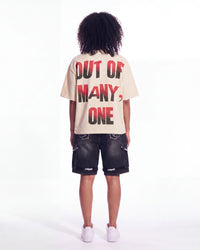 A child model shown from the back wearing a cream-colored t-shirt with bold red and black text reading 'OUT OF MANY, ONE' and dark denim shorts, against a plain white studio background.