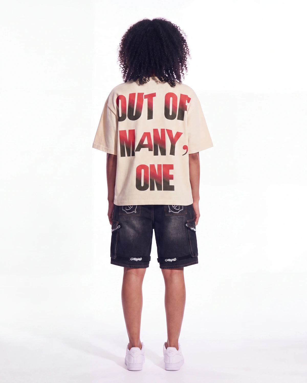 A child model shown from the back wearing a cream-colored t-shirt with bold red and black text reading 'OUT OF MANY, ONE' and dark denim shorts, against a plain white studio background.