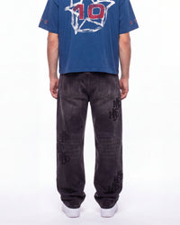 A person shown from the back wearing a blue graphic T-shirt with a large white and red '10' design, paired with dark gray washed jeans featuring black embroidered 'HO' logos on the legs, and white sneakers, against a plain white studio background.