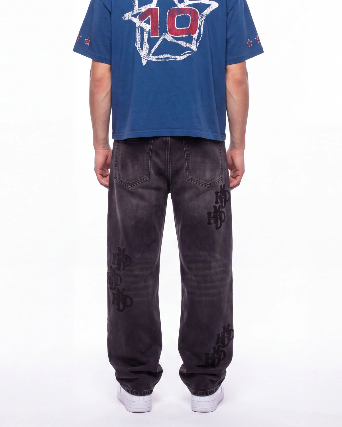 A person shown from the back wearing a blue graphic T-shirt with a large white and red '10' design, paired with dark gray washed jeans featuring black embroidered 'HO' logos on the legs, and white sneakers, against a plain white studio background.