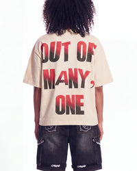 A person shown from the back wearing a cream-colored oversized t-shirt with the text 'OUT OF MANY, ONE' printed in a black-to-red gradient, paired with dark denim shorts.