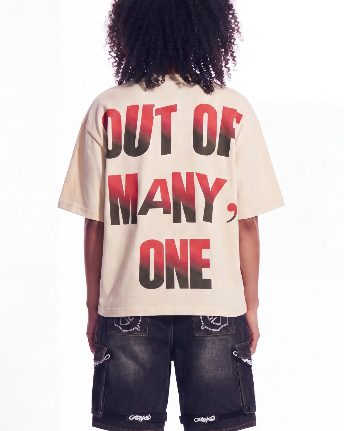 A person shown from the back wearing a cream-colored oversized t-shirt with the text 'OUT OF MANY, ONE' printed in a black-to-red gradient, paired with dark denim shorts.