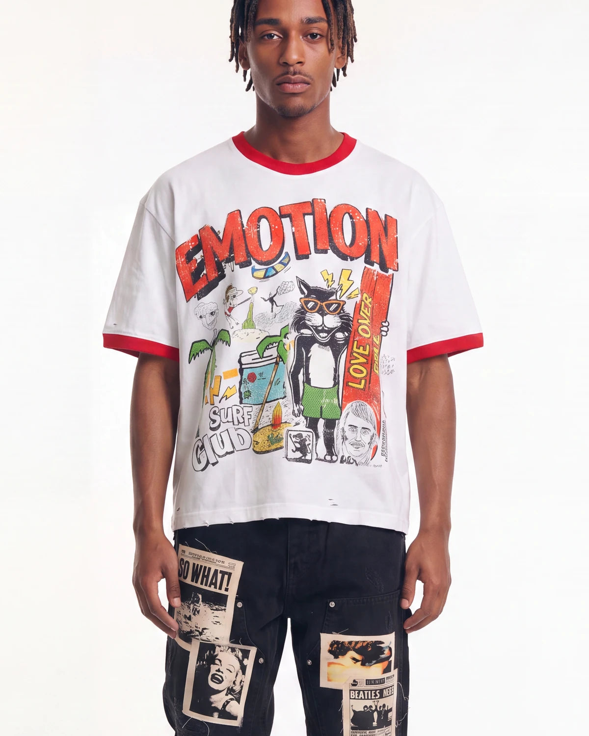 A man modeling a white graphic t-shirt with red trim and a colorful cartoon print featuring the word 'EMOTION' and a cat, paired with dark pants.