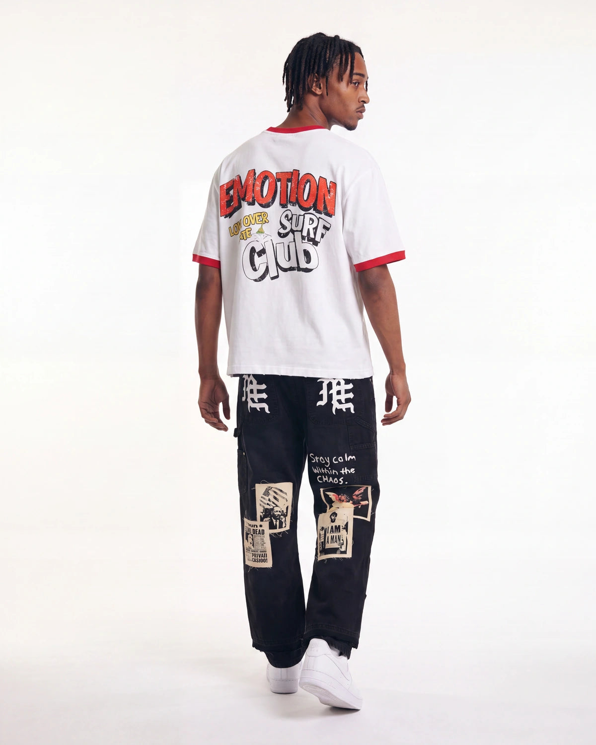 A model shown from the back wearing a white ringer t-shirt with red trim and a large graphic print on the back, paired with black graphic cargo pants and white sneakers, against a plain white studio background.