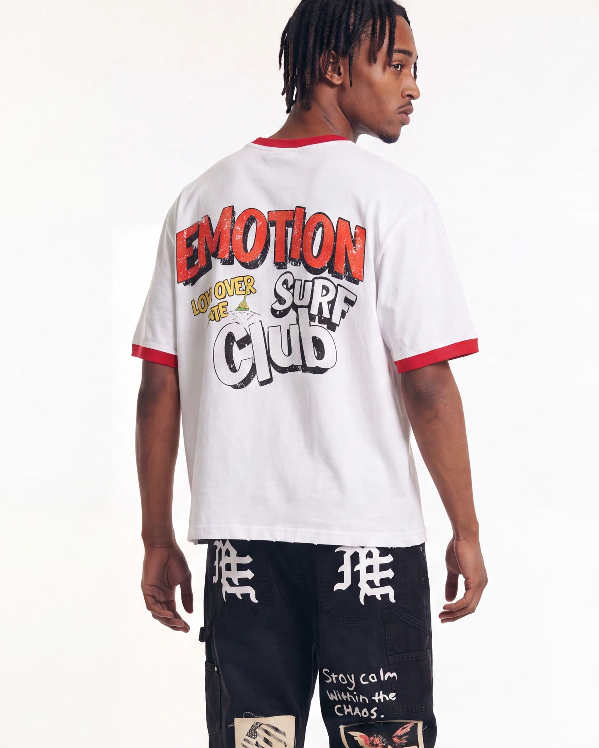 A man shown from the back wearing a white ringer t-shirt with red trim and a large graphic on the back that reads 'EMOTION SURF CLUB'. He is also wearing black graphic cargo pants.