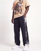 A model wearing dark navy track pants with white vertical 'VALLEY' and 'XSPORT' branding, paired with a beige graphic t-shirt and white sneakers, against a plain white studio background.