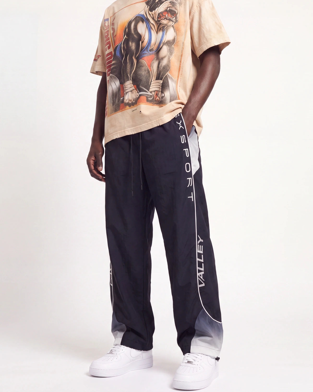 A model wearing dark navy track pants with white vertical 'VALLEY' and 'XSPORT' branding, paired with a beige graphic t-shirt and white sneakers, against a plain white studio background.