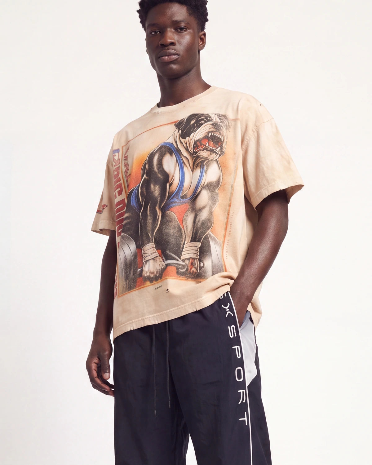 A male model wearing a beige graphic t-shirt with a bulldog weightlifting print and dark navy athletic pants with 'XSPORT' written vertically on the side, set against a plain white studio background.