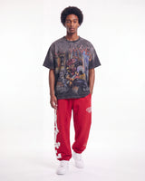 A model stands against a white studio background wearing a faded black graphic t-shirt, bright red sweatpants with white lettering, and white sneakers.
