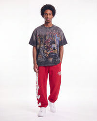 A model stands against a white studio background wearing a faded black graphic t-shirt, bright red sweatpants with white lettering, and white sneakers.