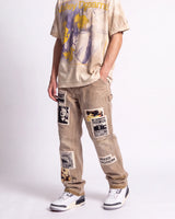 A person modeling beige cargo-style pants with multiple graphic patches and a matching oversized graphic T-shirt against a plain light gray studio background.