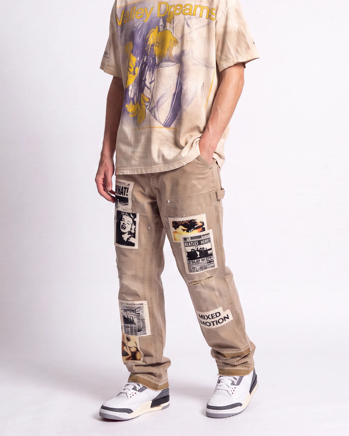 A person modeling beige cargo-style pants with multiple graphic patches and a matching oversized graphic T-shirt against a plain light gray studio background.