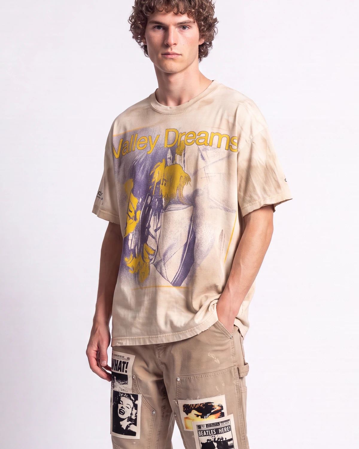 A man wearing a beige oversized graphic t-shirt with yellow and purple artwork and the text 'Violent Dreams' across the chest, paired with beige cargo pants featuring patch details, against a plain white studio background.