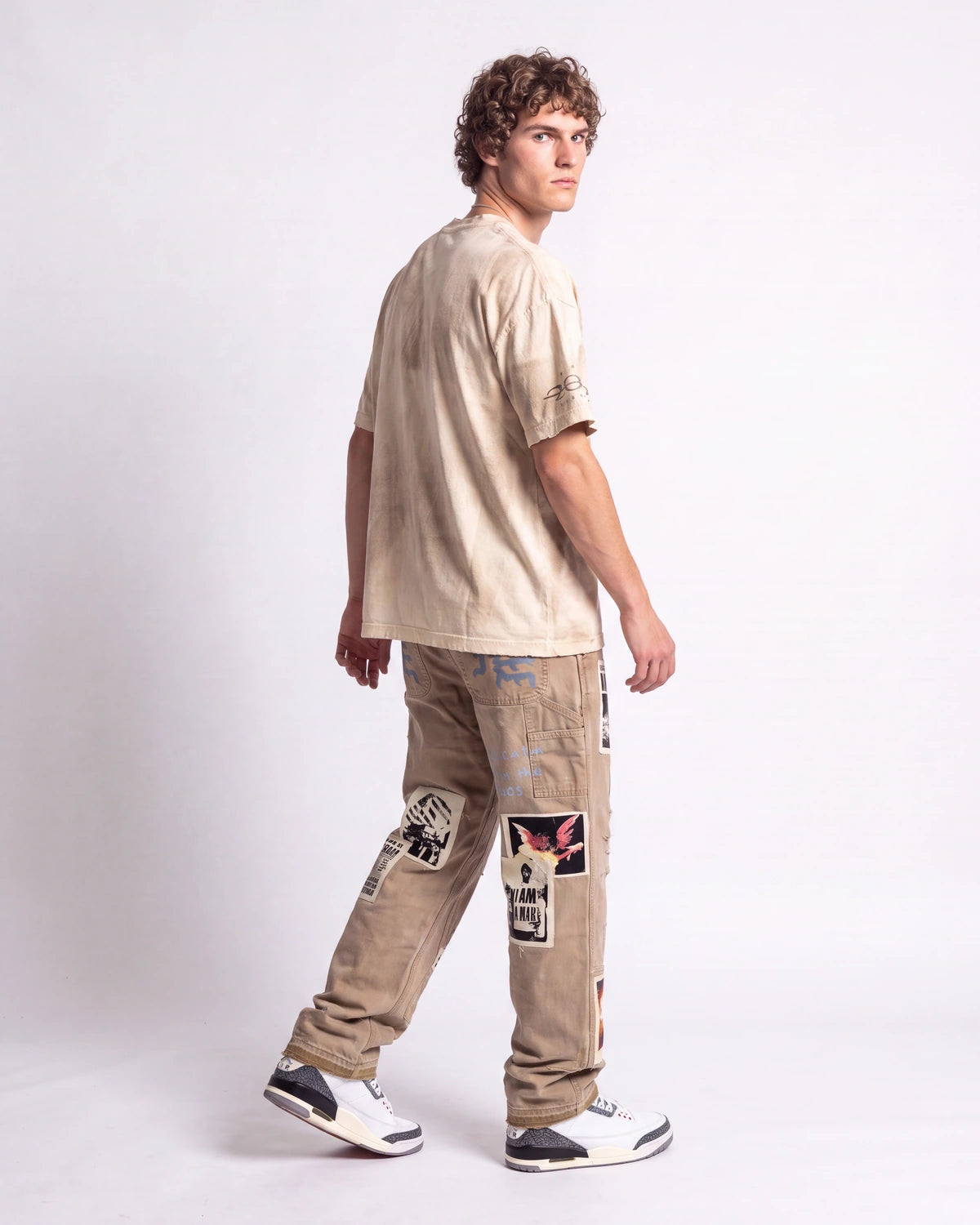A model shown from a rear three-quarter view wearing a beige t-shirt, beige patchwork cargo pants, and white sneakers with black accents, set against a plain light gray studio background.