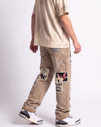 A person shown from the back wearing beige patchwork cargo pants with graphic prints, a light beige t-shirt, and white sneakers against a plain light gray studio background.