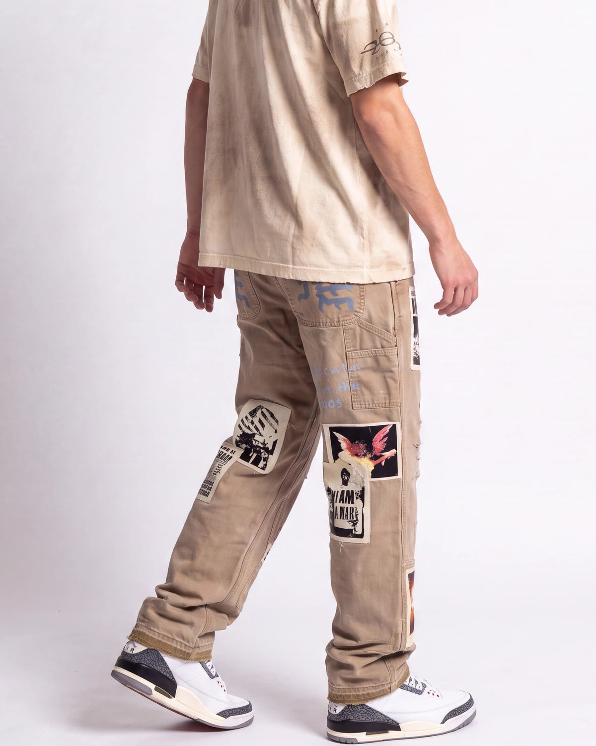 A person shown from the back wearing beige patchwork cargo pants with graphic prints, a light beige t-shirt, and white sneakers against a plain light gray studio background.