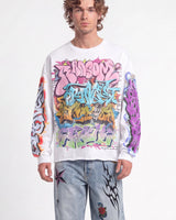 A man wearing a white crewneck sweatshirt with a large, colorful graffiti-style graphic print on the front and sleeves, paired with light-wash jeans featuring black and red graphic patches.