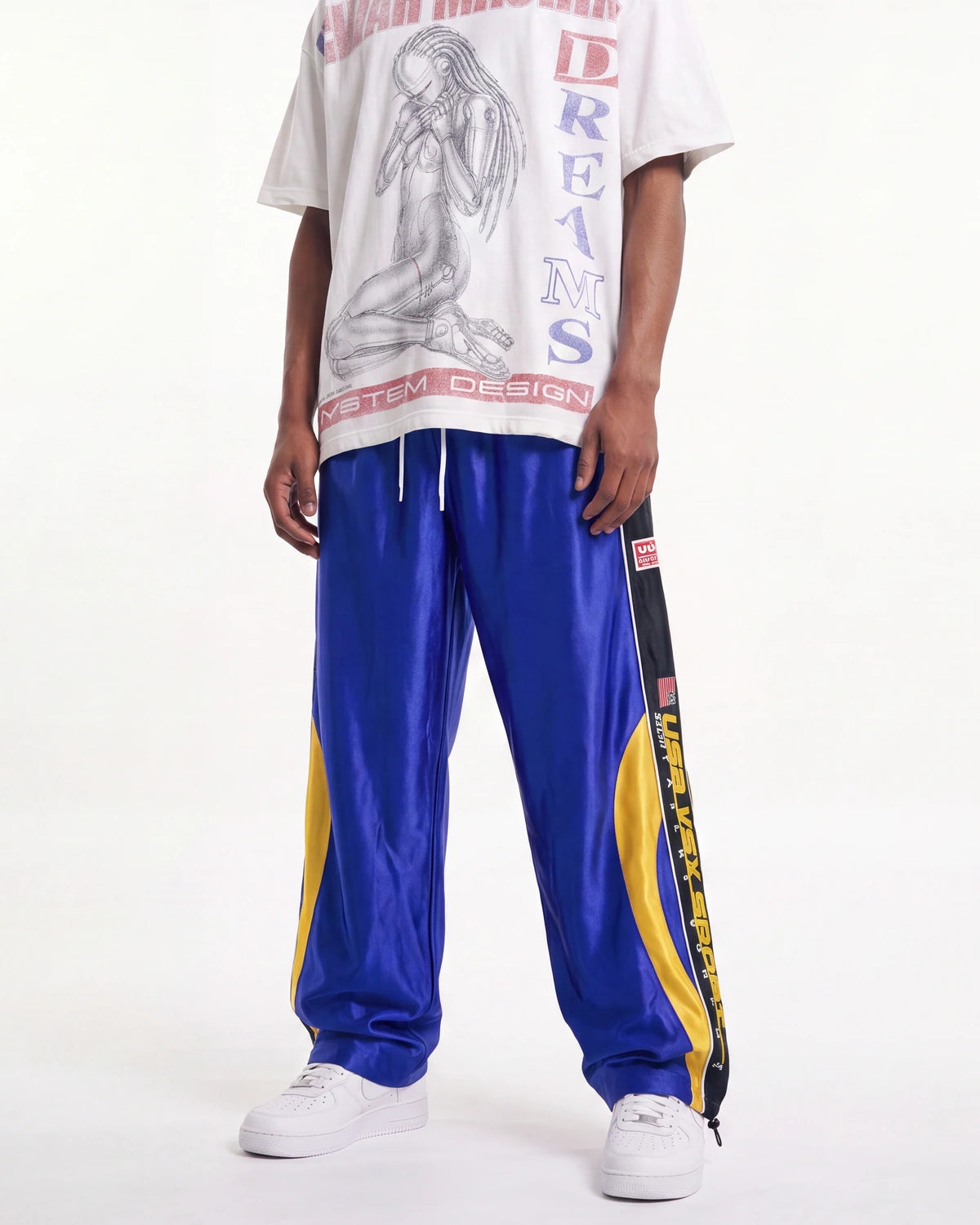 A model wearing a white graphic t-shirt and shiny royal blue sweatpants with yellow side stripes, paired with white sneakers, against a plain white studio background.