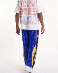 A person shown from the back wearing a white graphic T-shirt and shiny blue track pants with yellow and black side panels, against a plain light background.