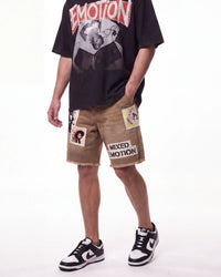 A person wearing a black graphic T-shirt with a large red and white 'EMOTION' print, tan distressed shorts with patch details and 'MIXED EMOTION' text, and black-and-white sneakers, photographed against a plain white studio background.