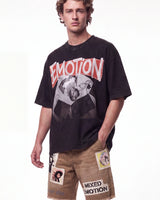 A male model wearing a black graphic t-shirt with a distressed red and white 'EMOTION' logo and a grayscale photo print, paired with tan shorts featuring patchwork details and 'MIXED EMOTION' text.