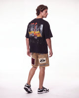 A man shown from the back wearing a black graphic t-shirt, beige shorts with patchwork details, and black-and-white sneakers, photographed against a seamless white studio background.