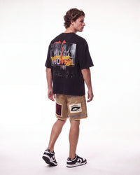 A man shown from the back wearing a black graphic t-shirt, beige shorts with patchwork details, and black-and-white sneakers, photographed against a seamless white studio background.
