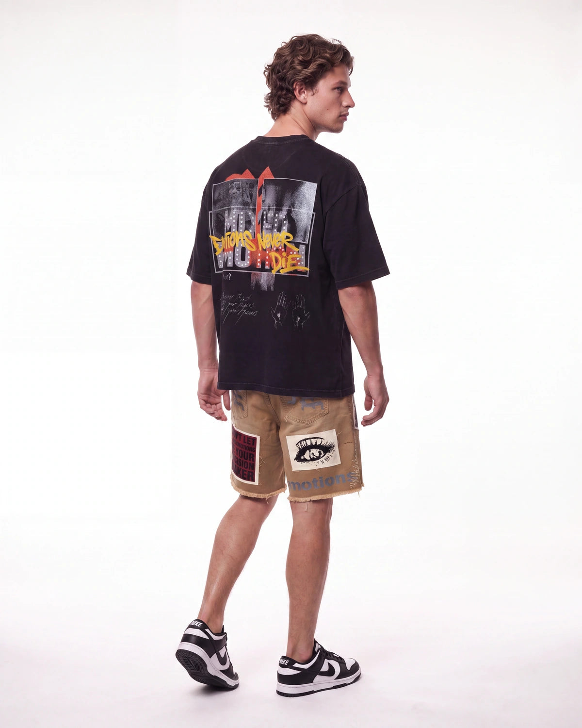 A man shown from the back wearing a black graphic t-shirt, beige shorts with patchwork details, and black-and-white sneakers, photographed against a seamless white studio background.