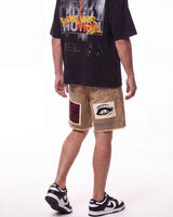 A person shown from the back wearing a black graphic T-shirt, tan distressed shorts with patch details, and black-and-white sneakers against a plain white studio background.