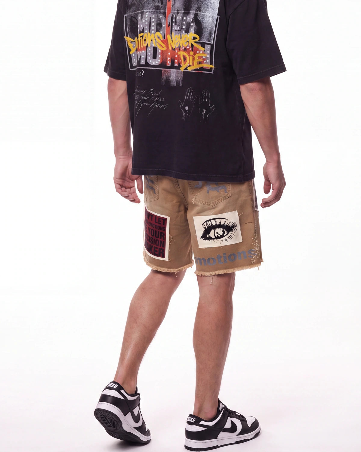 A person shown from the back wearing a black graphic T-shirt, tan distressed shorts with patch details, and black-and-white sneakers against a plain white studio background.