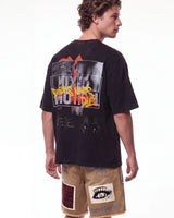 A man shown from the back wearing a faded black graphic t-shirt with a large 'This Is What We Do' design, paired with tan shorts, against a plain white background.