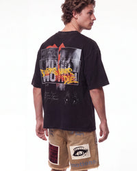 A man shown from the back wearing a faded black graphic t-shirt with a large 'This Is What We Do' design, paired with tan shorts, against a plain white background.