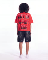 A child model shown from the back wearing a bright red oversized t-shirt with bold black and white text reading 'OUT OF MANY, ONE' and dark washed denim shorts, against a plain white studio background.