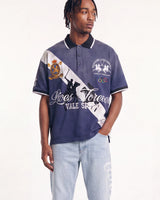 A man modeling a navy blue polo shirt with a diagonal white stripe, white graphics, and black trim, paired with light wash jeans against a plain white studio background.