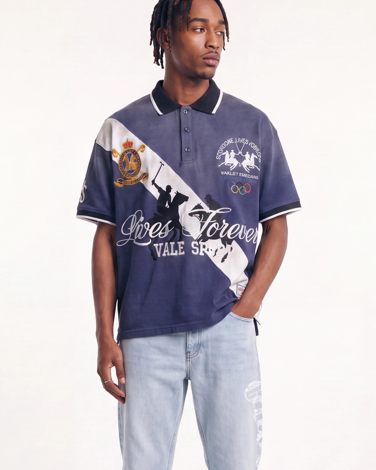 A man modeling a navy blue polo shirt with a diagonal white stripe, white graphics, and black trim, paired with light wash jeans against a plain white studio background.