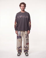 A male model stands against a plain white studio background wearing a dark gray long-sleeve t-shirt with 'VALE' printed across the chest and flame-patterned sleeves, paired with beige graphic sweatpants and black-and-white sneakers.