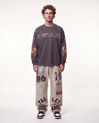 A male model stands against a plain white studio background wearing a dark gray long-sleeve t-shirt with 'VALE' printed across the chest and flame-patterned sleeves, paired with beige graphic sweatpants and black-and-white sneakers.
