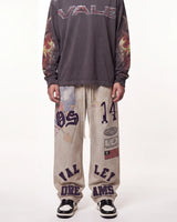A person wearing a dark gray long-sleeve sweatshirt with 'VALE' across the chest and flame graphics on the sleeves, paired with beige sweatpants featuring purple collegiate-style lettering and graphics, and black-and-white sneakers, against a plain white studio background.